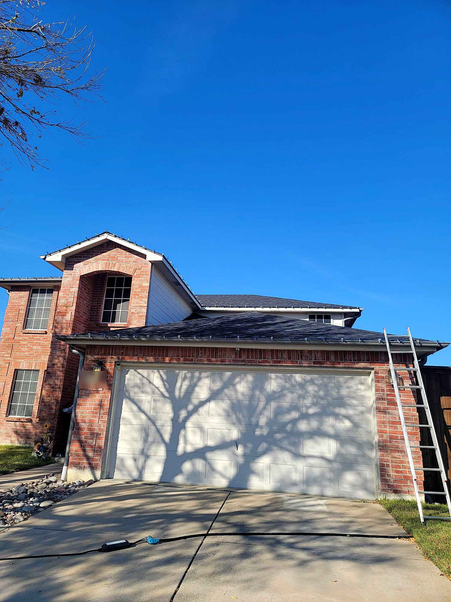 Daytime: a two-story brick house with light strands clipped along the roofline and a ladder against the garage